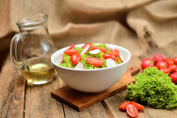 Green salad Lollo Biondo with tomatoes and radishes in a white bowl on wooden table, a pitcher of oil in the background