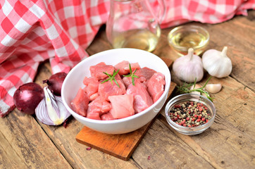 Close-up of raw pork meal diced in a bowl, garlic, half of onion, pepper, jug with oil and checkered red tablecloth in the background