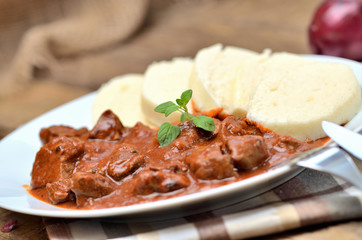 Close-up of pork goulash meat with dumplings on white plate, cutlery, garlic, onion, pepper, tablecloth in the background - typical Czech food