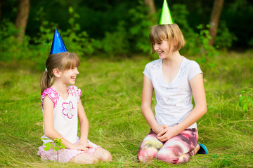 friendly children in festive cone caps, sit on grass, have fun together as celebrate birthday look with happy expressions at camera