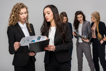 selective focus of beautiful businesswoman looking at charts and graphs while holding clipboard near colleague with attractive coworkers on background