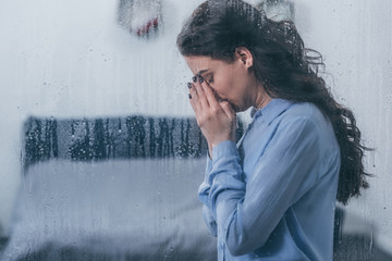 depressed woman covering face with hands and crying at home through window with raindrops and copy...