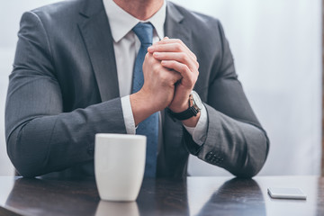 cropped view of man in gray suit sitting at wooden table with white cup and smartphone at home, grieving disorder concept