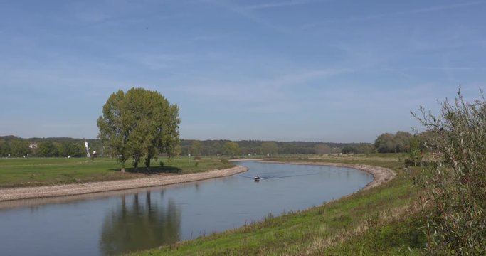 a small yacht sails upstream on the river - leaving a wake pattern in the undisturbed water. Veluwe moraine hill and highway A348 in background.