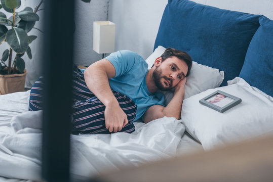 selective focus of sad man lying in bed near picture of woman on pillow at home