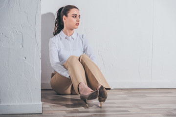 upset woman in white blouse and beige pants sitting on floor near white wall at home, grieving disorder concept