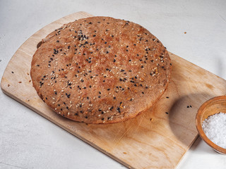 Round homemade bread made of dark flour sprinkled with sesame seeds lies on a wooden kitchen house. Coarse salt in a white salt shaker, linen napkin in the background.