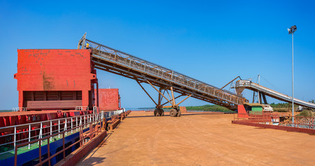 Conveyor belt loading bauxite aluminum ore into a capesize bulk carrier ship at a river bank jetee in the Kamsar, Guinea, West Africa.