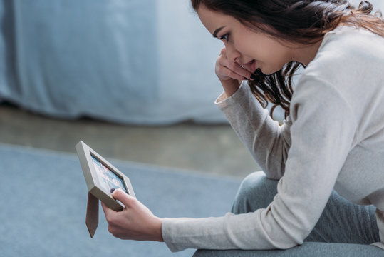 Selective Focus Of Upset Woman Looking At Picture Frame, Crying And Wiping Tears At Home