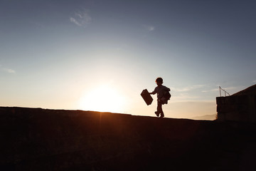 A little boy runs along a ledge at sunset with a suitcase in his hand. Little traveler. Tourism. Vintage. Escape from home. Silhouette.