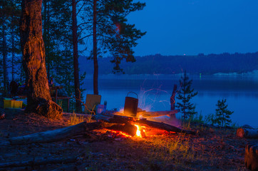Big night bonfire at the shore of the river in a forest glade, flames, sparks.