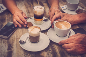 Close up aged caucasian hands with coffee cappuccino at the bar for breakfast - wooden table and vintage romantic color tones - mobile phone with the people - love and couple concept