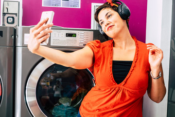 Cheerful beautiful caucasian young woman take selfie picture with the modern smart phone in a laudromat service store listening music with headphones and enjoying the wait for the service -