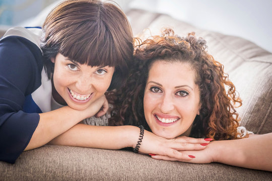 nice time and happiness with laugh and smiles for two caucasian female friends lay down together on the sofa at home. friendship concept for indoor picture with window natural light