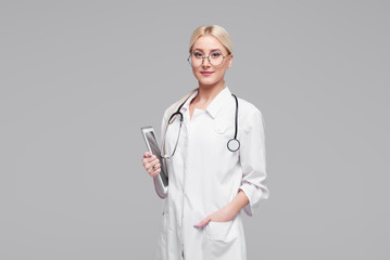 Young positive blond doctor woman in round glasses, in medical white coat with stethoscope taking notes on tablet pc. Isolated grey background