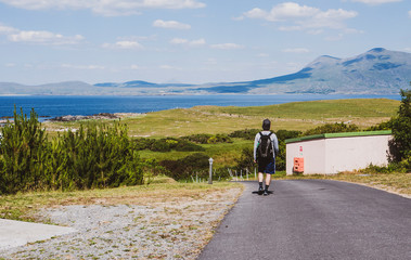 Man with a backpack, cap & shorts walking down a hill, with mountains & ocean in the distance. Taken in summer in Renvyle, along the Wild Atlantic Way in Ireland.