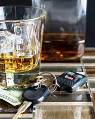 Glass of alcohol with ice cubes.  Money and keys in the foreground with a nearly empty bottle in the background.  Fading into black background.  Suggestive of drinking and driving or alcohol abuse.
