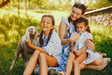 three beautiful and cute girls in blue dresses with beautiful hairstyles and make-up sitting in a sunny green garden and playing with a goat