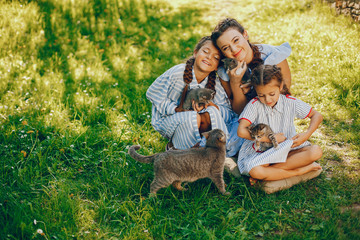 Naklejka na ściany i meble three beautiful and cute girls in blue dresses with beautiful hairstyles and make-up sitting in a sunny green garden and playing with a cats