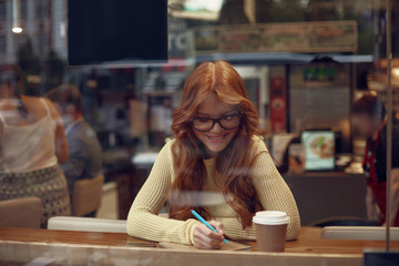 Charming red-haired girl writing on spiral notebook in cafe