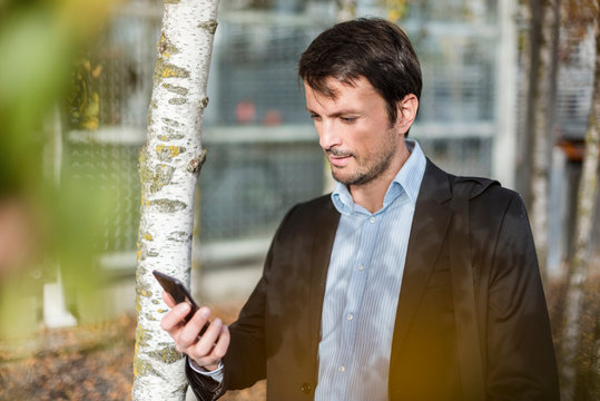 Portrait of a businessman under trees, using smartphone