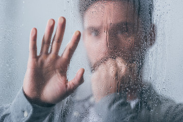 upset man covering mouth with hand and touching window with raindrops