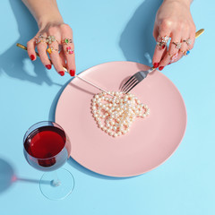 Female hands cut pearl beads with a knife and fork in a pink plate on a blue table. Creative concept. Top view.