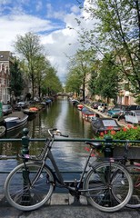 Fototapeta premium Photo showing the bridge on one of the Amsterdam canals. On the bridge parked bicycle next to bicycles flowerpots with beautiful red flowers. Old wooden boats parked along the canal