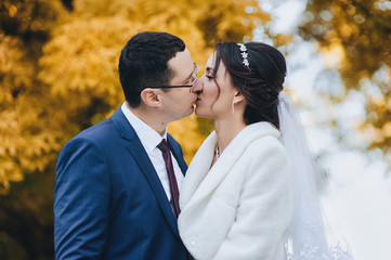 Beautiful and smiling newlyweds kiss tenderly, in autumn in a park with yellow leaves. Closeup portrait of stylish groom in glasses and cute bride in white dress. Wedding photography.