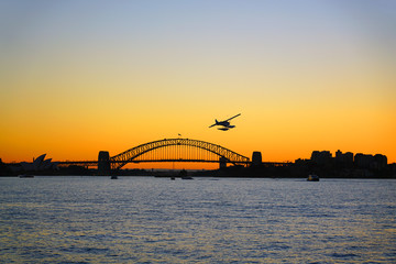 Sunset view of a seaplane flying in the orange sky by the iconic steel Sydney Harbour Bridge in New South Wales, Australia