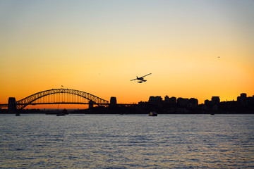 Naklejka premium Sunset view of a seaplane flying in the orange sky by the iconic steel Sydney Harbour Bridge in New South Wales, Australia
