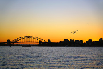 Sunset view of a seaplane flying in the orange sky by the iconic steel Sydney Harbour Bridge in New South Wales, Australia