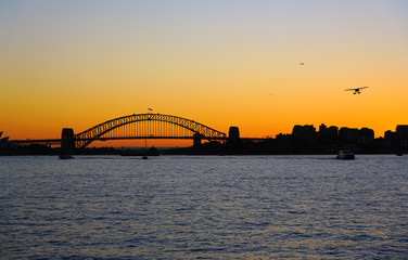 Sunset view of a seaplane flying in the orange sky by the iconic steel Sydney Harbour Bridge in New South Wales, Australia