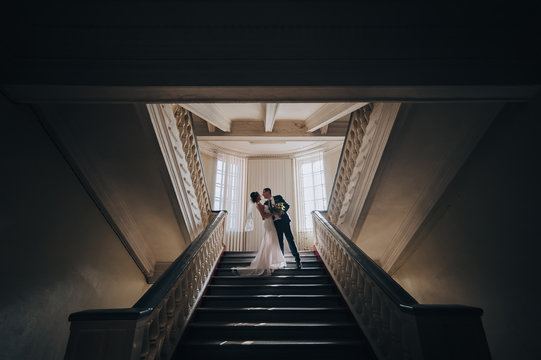 Newlyweds in love are standing on the stairs in a dark building with a bright window. Portrait of a stylish groom with glasses and a beautiful cute bride in a white dress. Wedding photography.