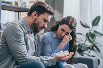 woman sitting and crying on couch while man holding baby shoes in room, grieving disorder concept