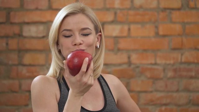 A close-up of a charming blonde in dark tank top standing against the brick wall and biting a big red apple and chewing it
