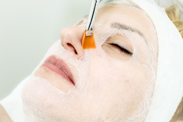 beautician worker holding paintbrush  applying facial cosmetic serum  in biological treatment to female client  in beauty salon, eyes closed, close up 