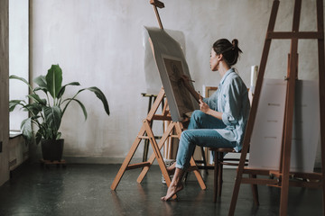 Female artist in spacious art studio sitting barefooted and drawing