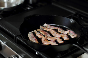 Strips of bacon frying in a cast iron pan on the stove.