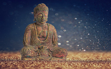 A Buddha meditating surrounded by sand and a blue bokeh background.