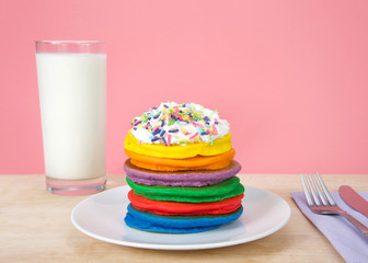 Stack of bright colorful pancakes topped with whipped cream and candy sprinkles, utensils and glass...