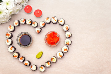 Romantic heart sushi dinner. Maki rolls with ginger, wasabi and soy sauce, candles and flowers. Concept for Valentine's Day or March 8th. Stone background, top view.