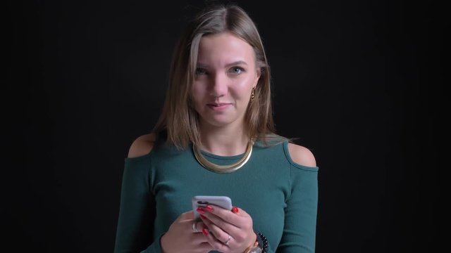 Portrait of young brunette caucasian girl positively nodding into camera and watching into smartphone on black background.