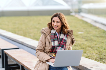 Smiling Caucasian brunette in jacket sitting on bench outside and using laptop.