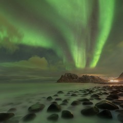 Northern lights in Lofoten islands, Norway. Green Aurora borealis. Starry sky with polar lights. Night winter landscape with aurora, sea with sky reflection, rocks, beach and snowy mountains. 