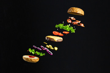 Floating burger isolated on black wooden background. Ingredients of a delicious burger with ground beef patty, lettuce, bacon, onions, tomatoes and cucumbers - obrazy, fototapety, plakaty