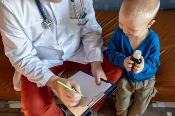 Home doctor in white coat, cap and glasses with gray beard examines two children teenagers at home....