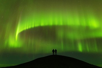 Naklejka na ściany i meble Aurora borealis and silhouette of standing lovers. Jokulsarlon, Iceland. Aurora and happy man. Sky with stars and green polar lights. Night landscape with aurora and people. Nature background concept