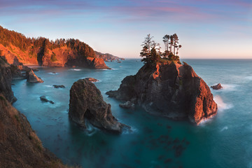 Sunset at Natural Bridges along Samuel H. Boardman State Scenic Corridor, Oregon during a golden hour - sunbeams through trees with dense vegetation. Beautiful seascape with rocks. West Coast USA