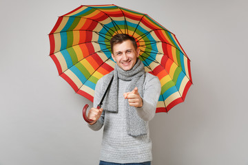 Handsome young man in gray sweater, scarf holding colorful umbrella pointing index finger on camera...